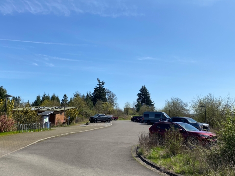 Image of an asphalt driveway with vehicles parked on either side and a brown building with a slanted roof to the left of the driveway.