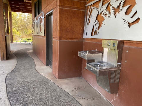 Image of a silver water fountain and bottle filling station attached to a brown building with gray metal nature art panel above and concrete pathway below.