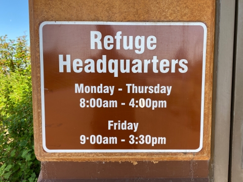 A brown sign with white letters indicating the operating hours of the refuge headquarters office.