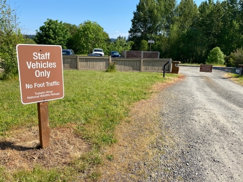 Image of a brown sign with white lettering designating a staff parking area. A gravel road and gate are next to the sign.