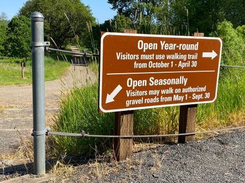 Image of a brown sign with white lettering and arrows directing toward trails. The sign is surrounded by a metal fence, gravel and green grass.