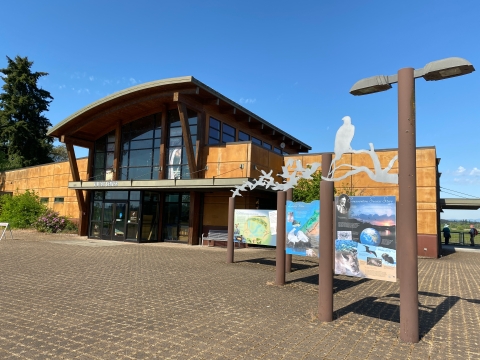 image of three colorful signs with brown posts and sliver metal silhouettes of birds above them. The signs are on a concrete paver plaza. A light brown building and blue sky is in the background.