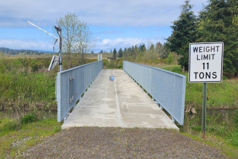 Image of a cream colored concrete bridge with gray railings with a white weight limit sign and water, grass, and trees on either side.