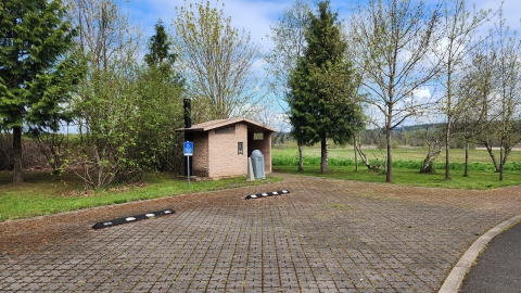 Image of a small light brown bathroom building with parking spots and trees surrounding it.