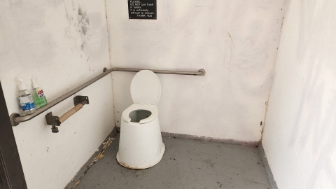 Image of a pit toilet interior with a raised white seat and grab bars to the left and behind the seat.