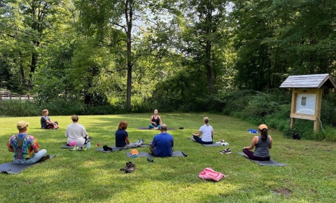 people sit cross-legged on mats on green grass with green trees in the background and a wooden information kiosk on the right