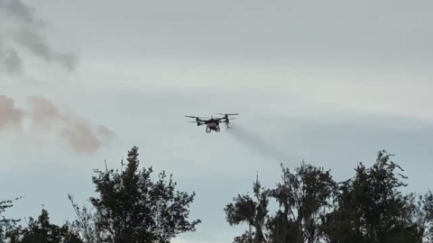 A drone flies in the sky, with a cloud of herbicide mist trailing behind it.