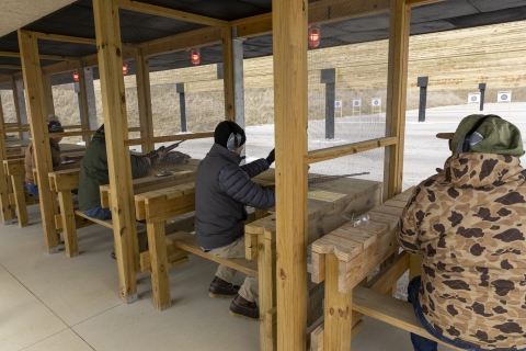 People sitting on wooden benches holding firearms and shooting at a range