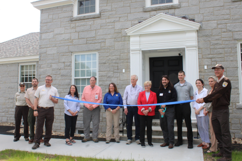 15 people gather in front of a stone house. On either end of the group a uniformed USFWS employee holds a blue ribbon.