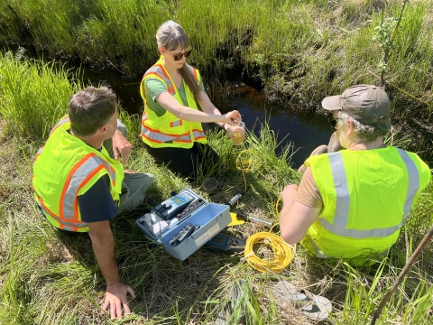 Three people sitting in the grass next to a stream holding small water quality instruments. One person is holding one of the tools while the other two people watch her. 