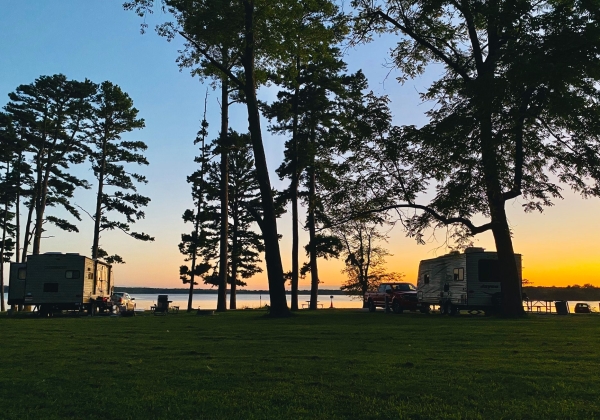 Campers set up, surrounded by evergreen trees with a lake and yellow sunset in the background. 
