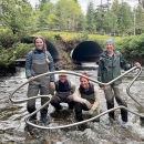4 people sanding downstream of undersized road culverts in a stream holding a big metal fish