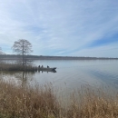 A calm lake landscape with a small boat up against a marshy shoreline with trees. A net held up with buoys is visible in a line extending out into the lake from the boat