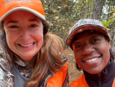 Two women wearing orange and camo, smile together in the woods