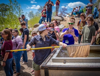 A high school student holding a net over a fish trailer. There are hundreds of people in the background as well as green trees and a blue sky with clouds.