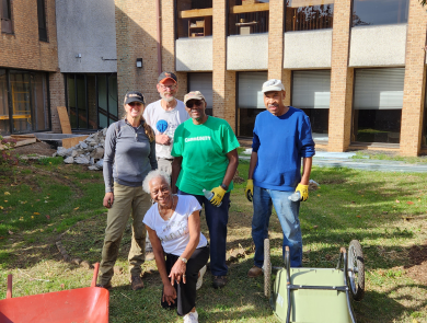 Five people stand in the yard of a building with wheelbarrows and other gardening tools