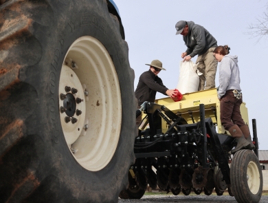 Three people standing on a tractor-pulled seed planter, with a large tractor tire in the foreground.