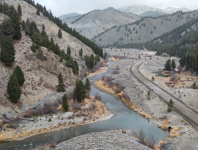 An aerial view over a winding river with a road along one side and mountains with evergreen trees on the other. there is a light dusting of snow on the road and terrain.