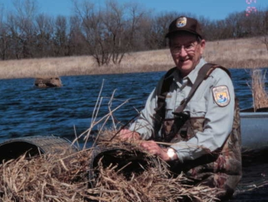Biologist wearing waders and sitting in a boat on the water fixing a grass/straw hen house that sits on top of the water.