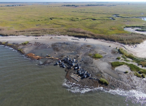 In this aerial image, Gandys Beach is visible in the foreground, as are a handful of volunteers and project staff and large square, gray containers filled with reef materials. A green marsh is visible behind the beach.