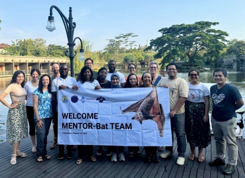 A group of about 18 people stand smiling at the camera holding a large banner that reads "Welcome MENTOR-Bat Team." 