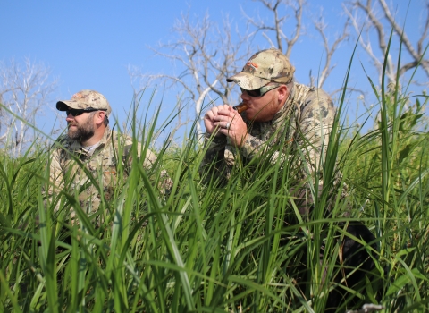 Two men dressed in camouflage sitting in grass. One man is holding a duck call to has mouth.