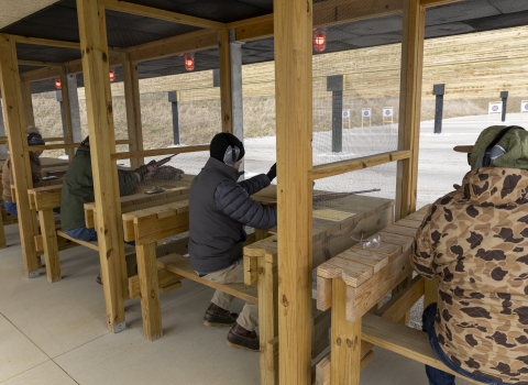 People sitting on wooden benches holding firearms and shooting at a range